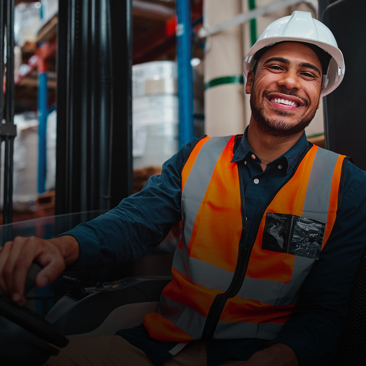 A man in a hard hat and vest sits in a forklift, smiling.